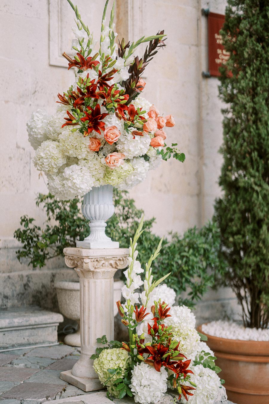 Elegant floral arrangement featuring white hydrangeas, red lilies, and peach roses in an ornate stone urn, set against a rustic stone wall backdrop. Perfect for weddings or outdoor events.