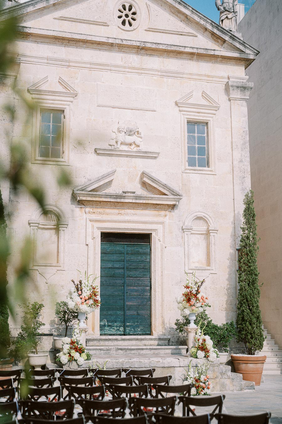 Historic stone building with wooden doors prepared for an outdoor wedding ceremony, featuring rows of black chairs and elegant floral arrangements in shades of white, peach, and red.