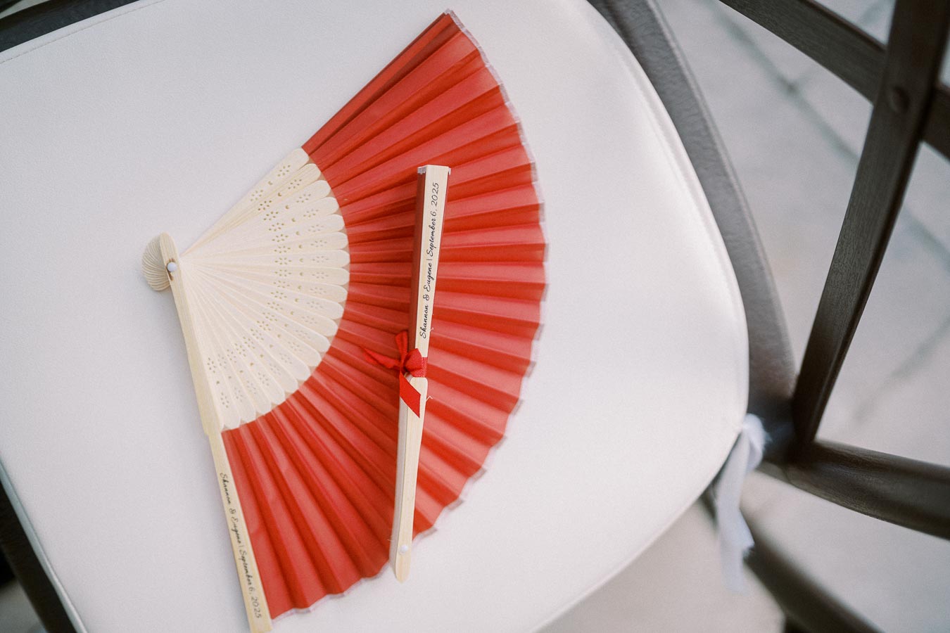 Red and beige folding hand fan with personalized inscription lying on a white chair.