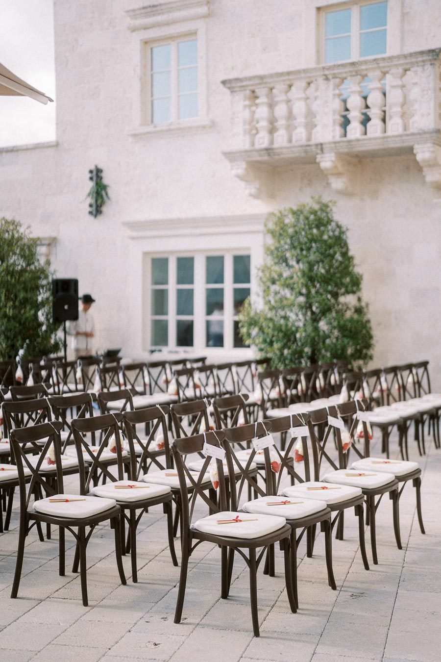 Outdoor wedding ceremony setup with rows of elegant wooden chairs, each adorned with white cushions and tied with small tags, set against a backdrop of a beautiful stone building and greenery.