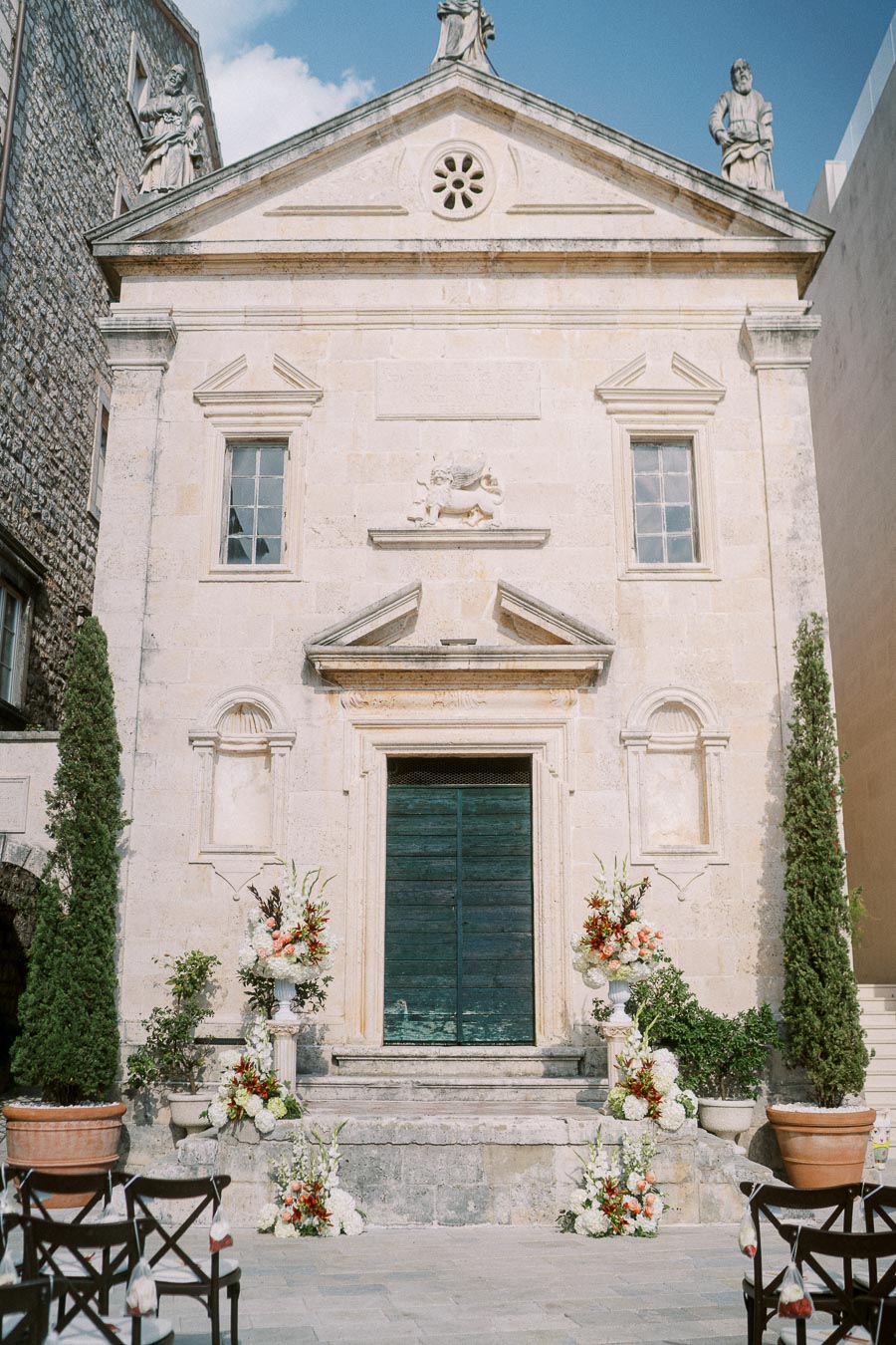 Historic stone building with ornate architecture, decorated with large floral arrangements and surrounded by tall potted plants under a clear blue sky.
