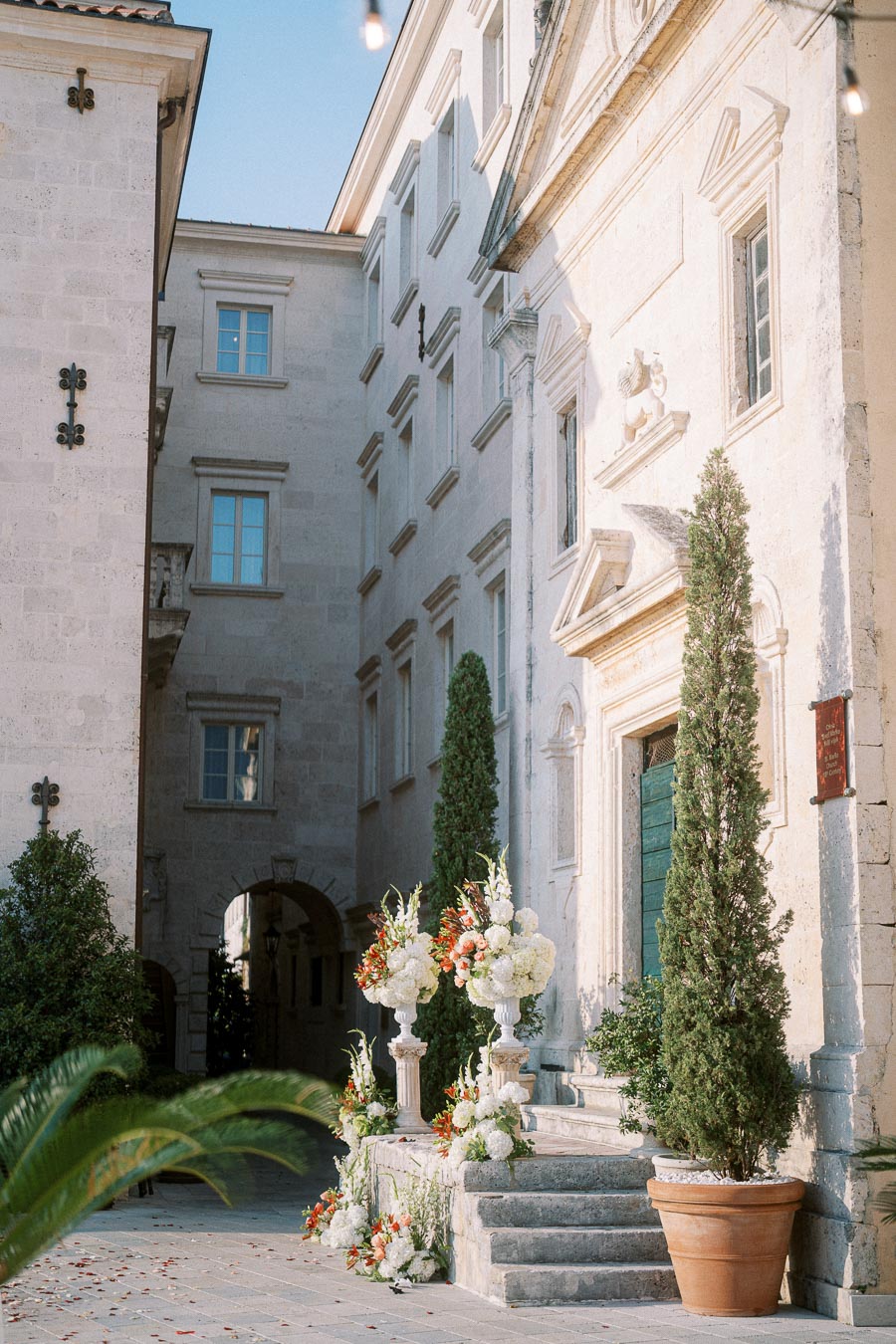 Elegant European courtyard with classic architecture featuring arched windows and decorative elements. A stairway adorned with white and orange floral arrangements leads to a large green door, framed by tall potted trees.
