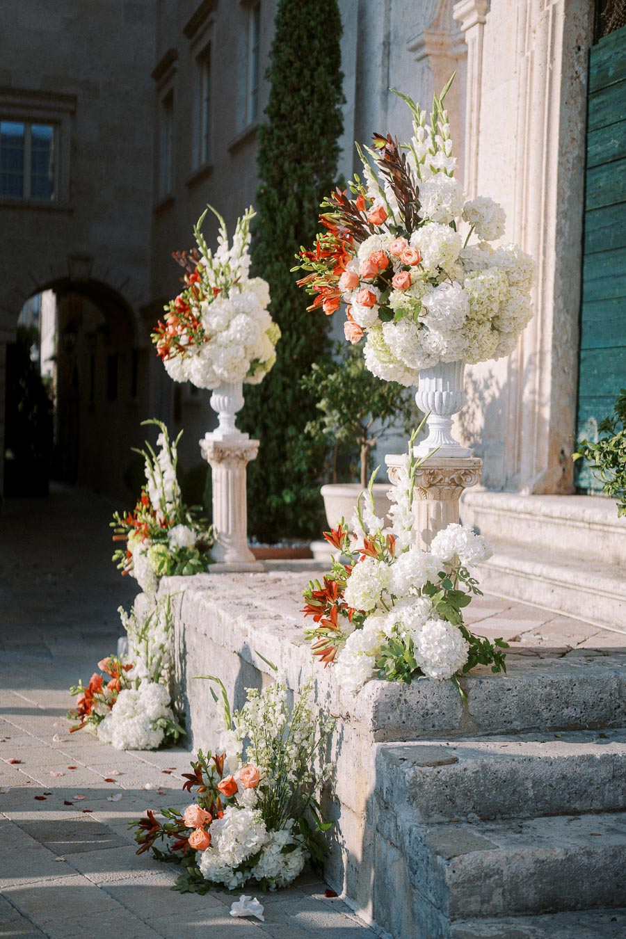 Luxurious outdoor wedding floral arrangements featuring white hydrangeas, orange roses, and greenery on elegant pedestals accenting stone steps at a classic venue.