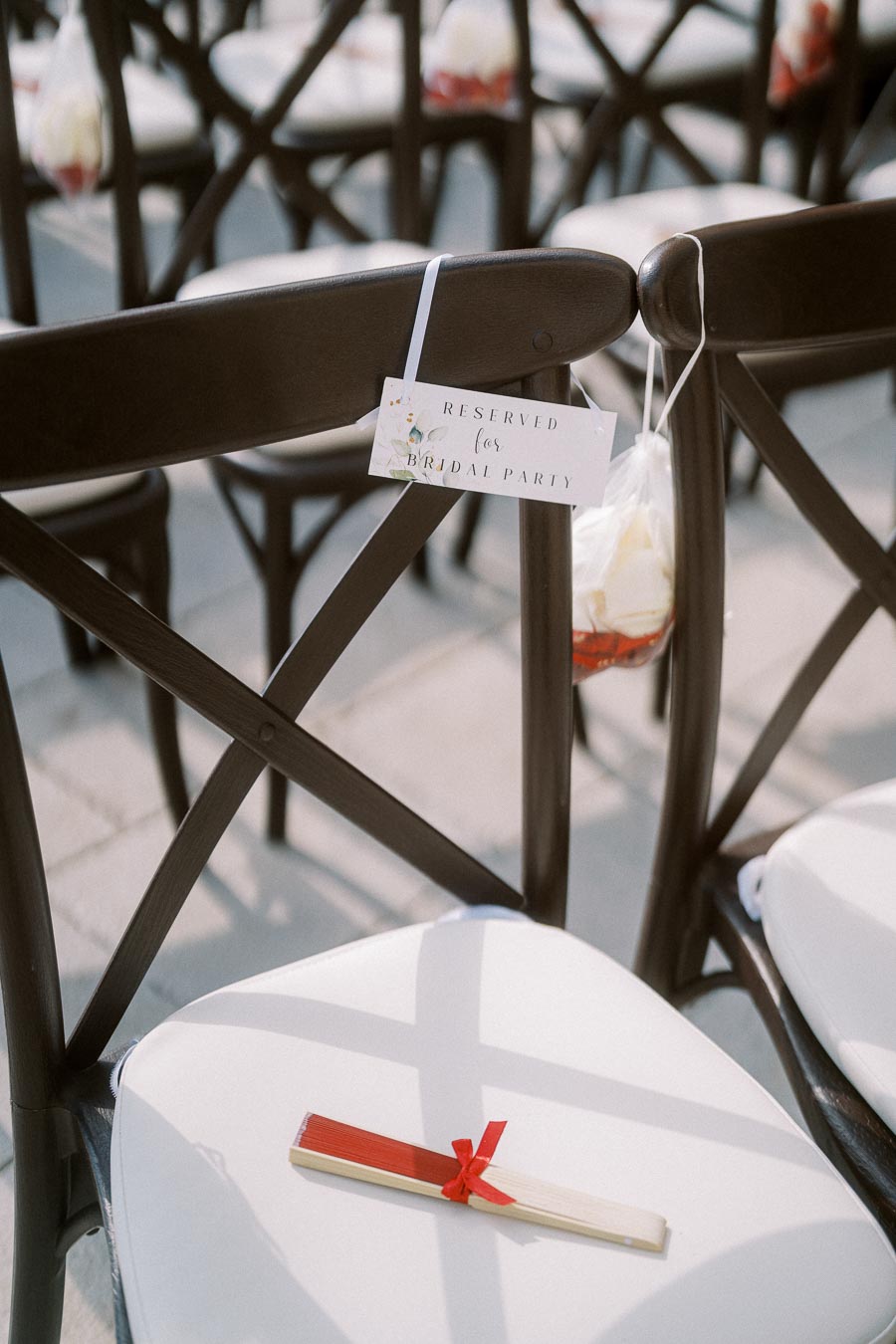 A row of elegant wooden chairs with white cushions set up for a wedding, featuring a Reserved for Bridal Party sign and a small fan tied with a red ribbon placed on one chair, creating a sophisticated atmosphere.