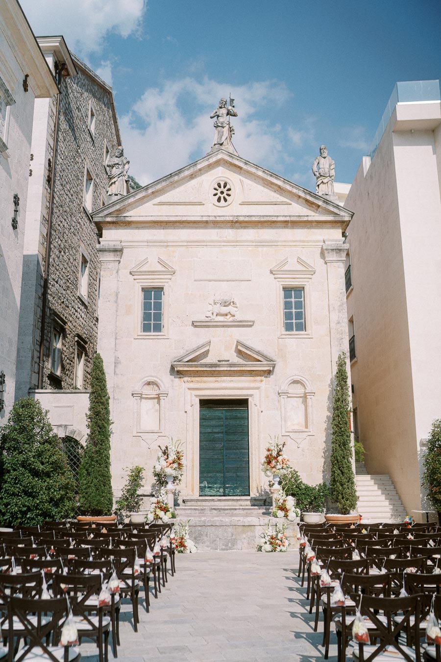 Historic stone church facade with decorative architecture, surrounded by greenery, set up for an outdoor wedding ceremony with rows of chairs and floral arrangements under a clear blue sky.