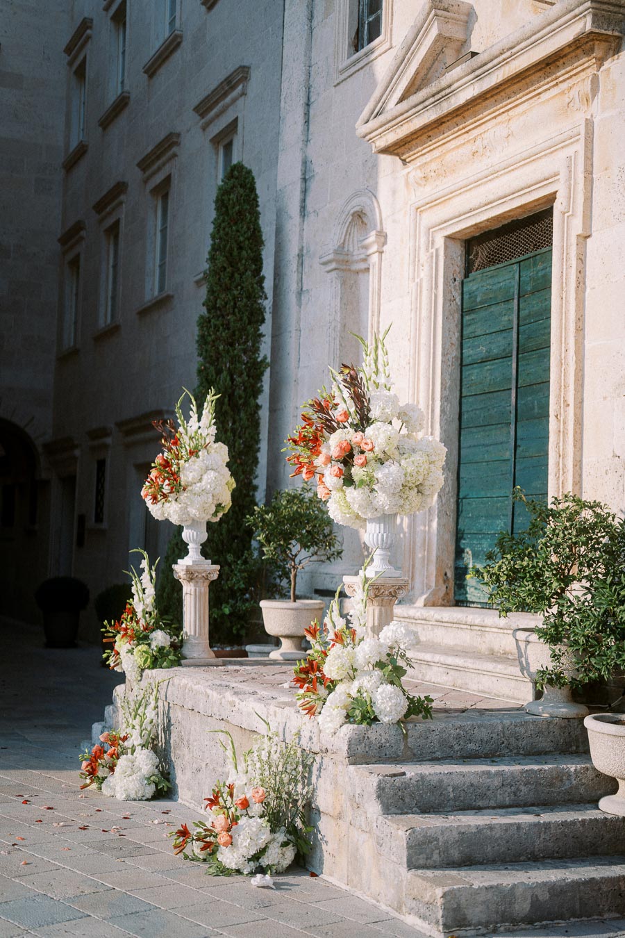 Elegant wedding venue decorated with white and red floral arrangements on stone steps, featuring vintage architecture and green doors in a picturesque outdoor setting.