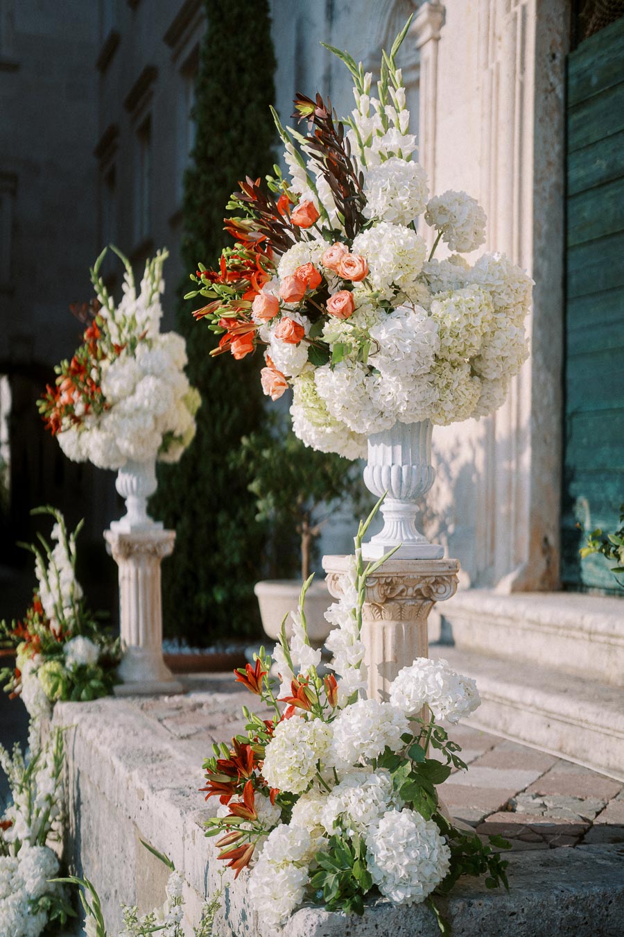 Elegant outdoor wedding floral arrangements featuring white hydrangeas and orange roses in ornate white vases on stone steps.