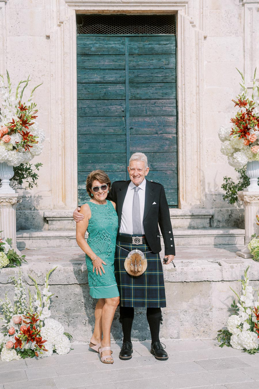 Elderly couple smiling and posing in front of an ornate, rustic doorway. The man is wearing a traditional Scottish kilt with a sporran, while the woman is dressed in a stylish turquoise dress. They are surrounded by large floral arrangements of white and red blooms, set against a stone staircase.