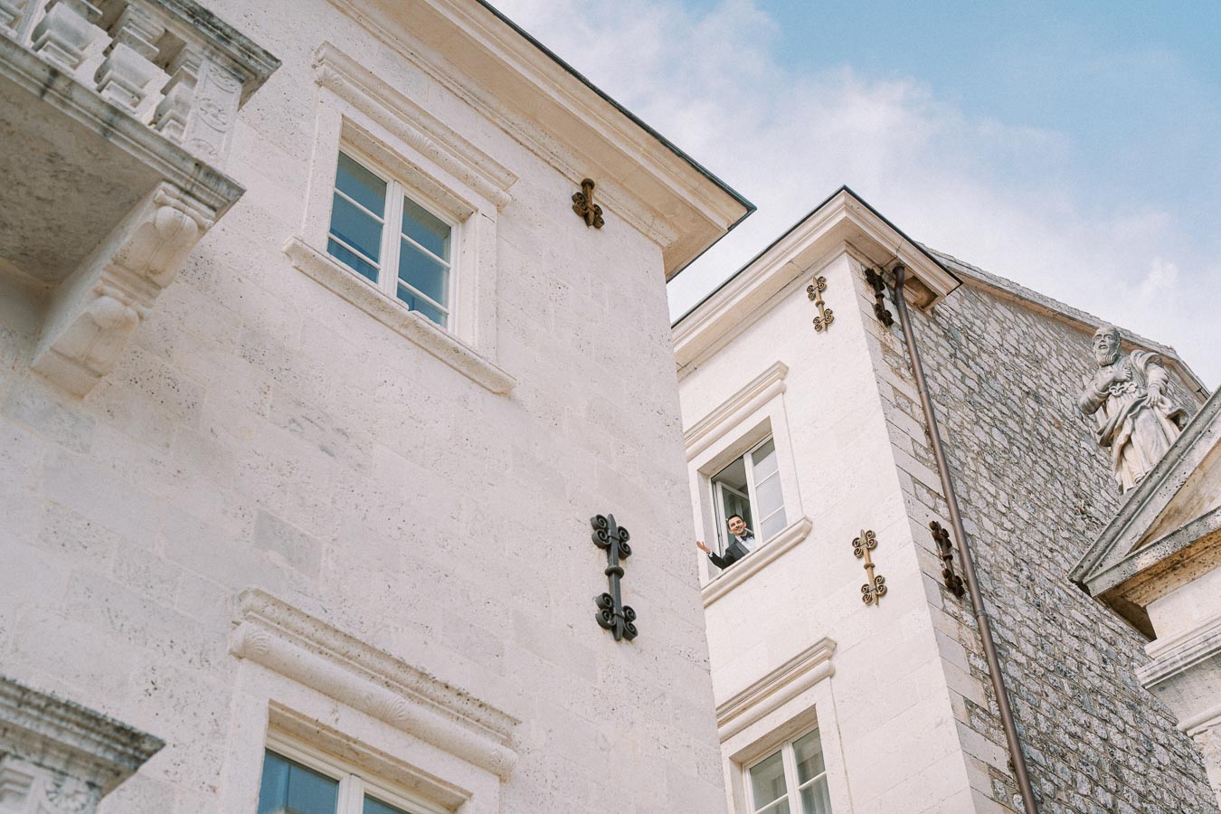 Elegant European architecture with a person waving from a window, featuring ornate details on light stone buildings against a clear blue sky.