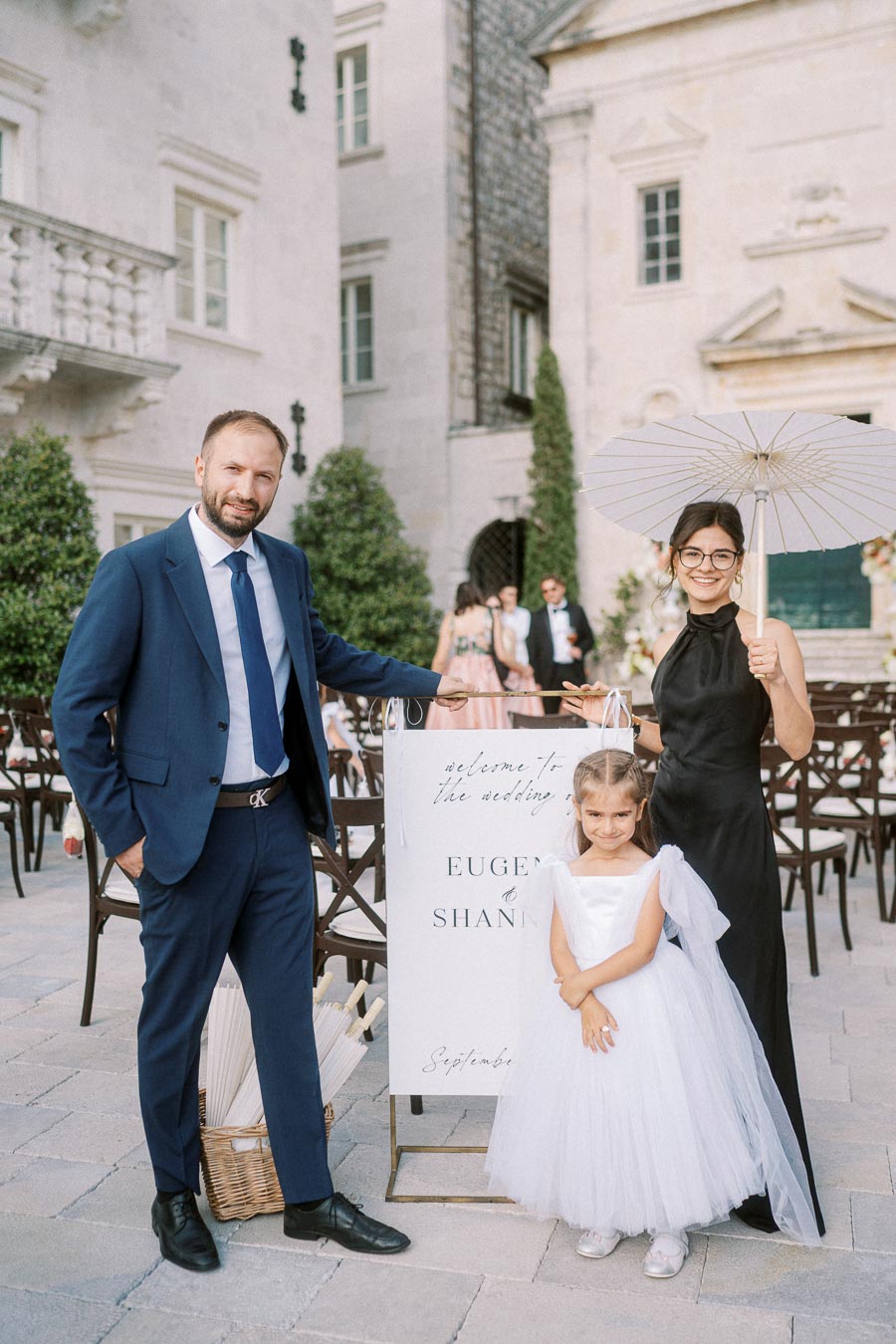 A man in a blue suit, a woman in a black dress holding a parasol, and a young girl in a white dress pose happily by a wedding welcome sign in an elegant outdoor venue.
