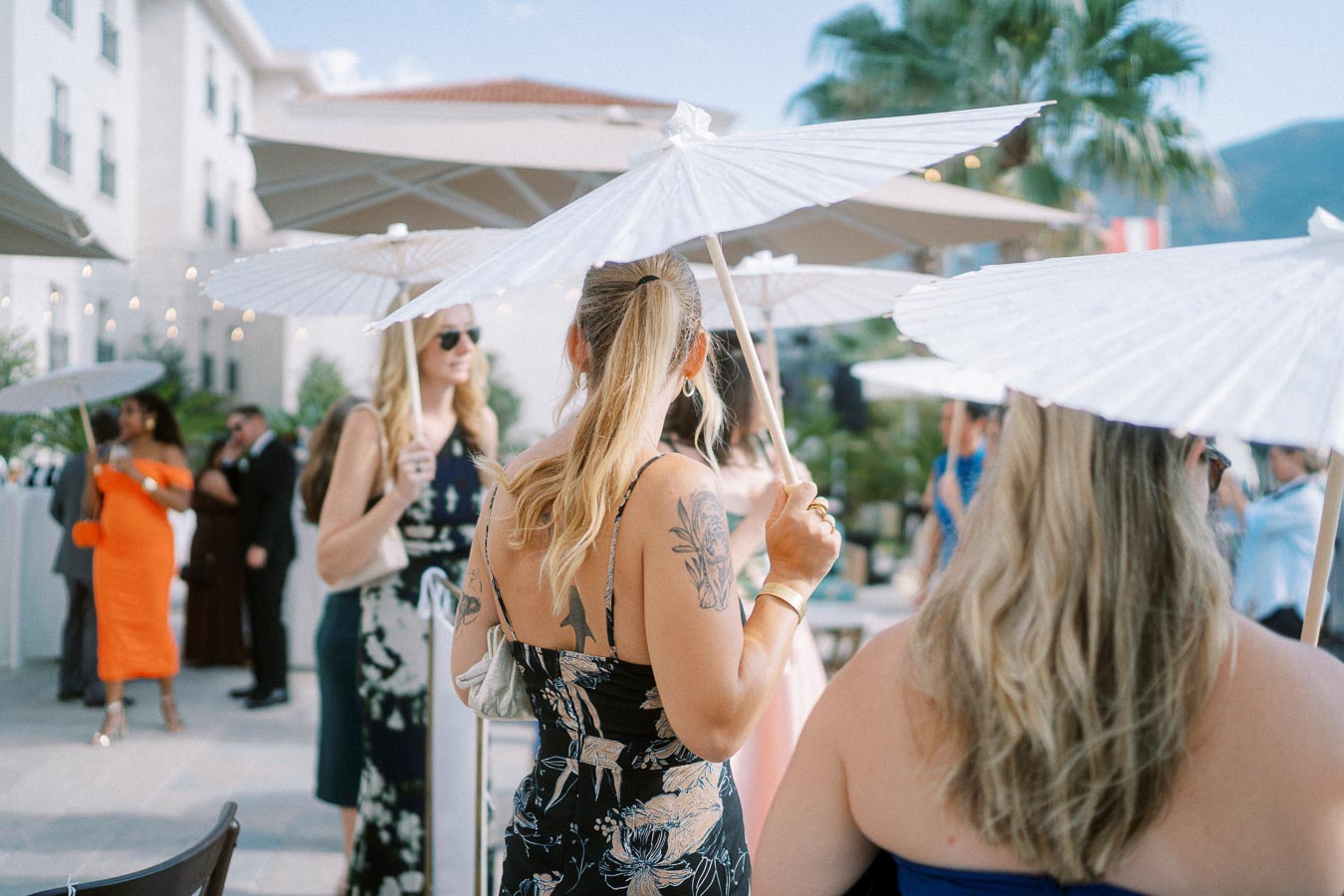 Outdoor gathering with people holding white umbrellas, dressed in summer attire, at a sunny venue with elegant decor and palm trees.