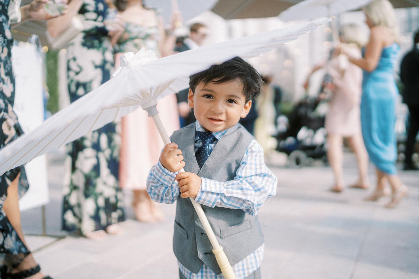 A young boy in a stylish gray vest and checkered shirt holds a white parasol at a formal outdoor event, surrounded by elegantly dressed people.