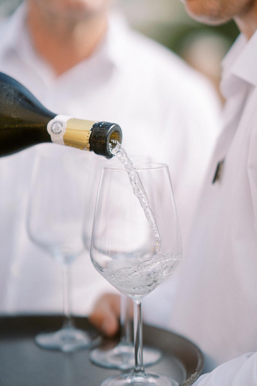 Bottle pouring sparkling wine into glasses at an event with people in white shirts.
