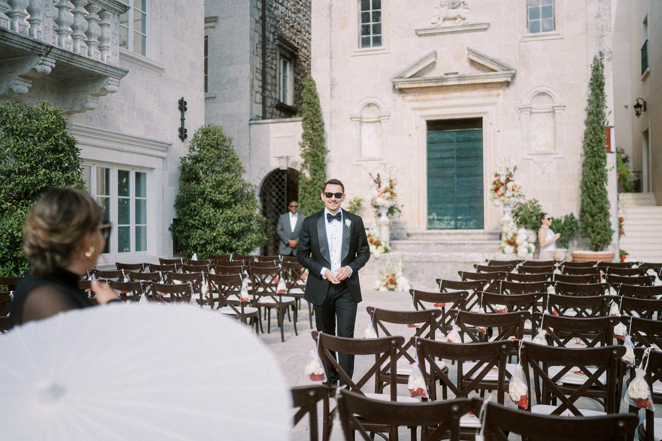 Groom in a tuxedo walking down the aisle at an elegant outdoor wedding venue with decorative floral arrangements and empty chairs.