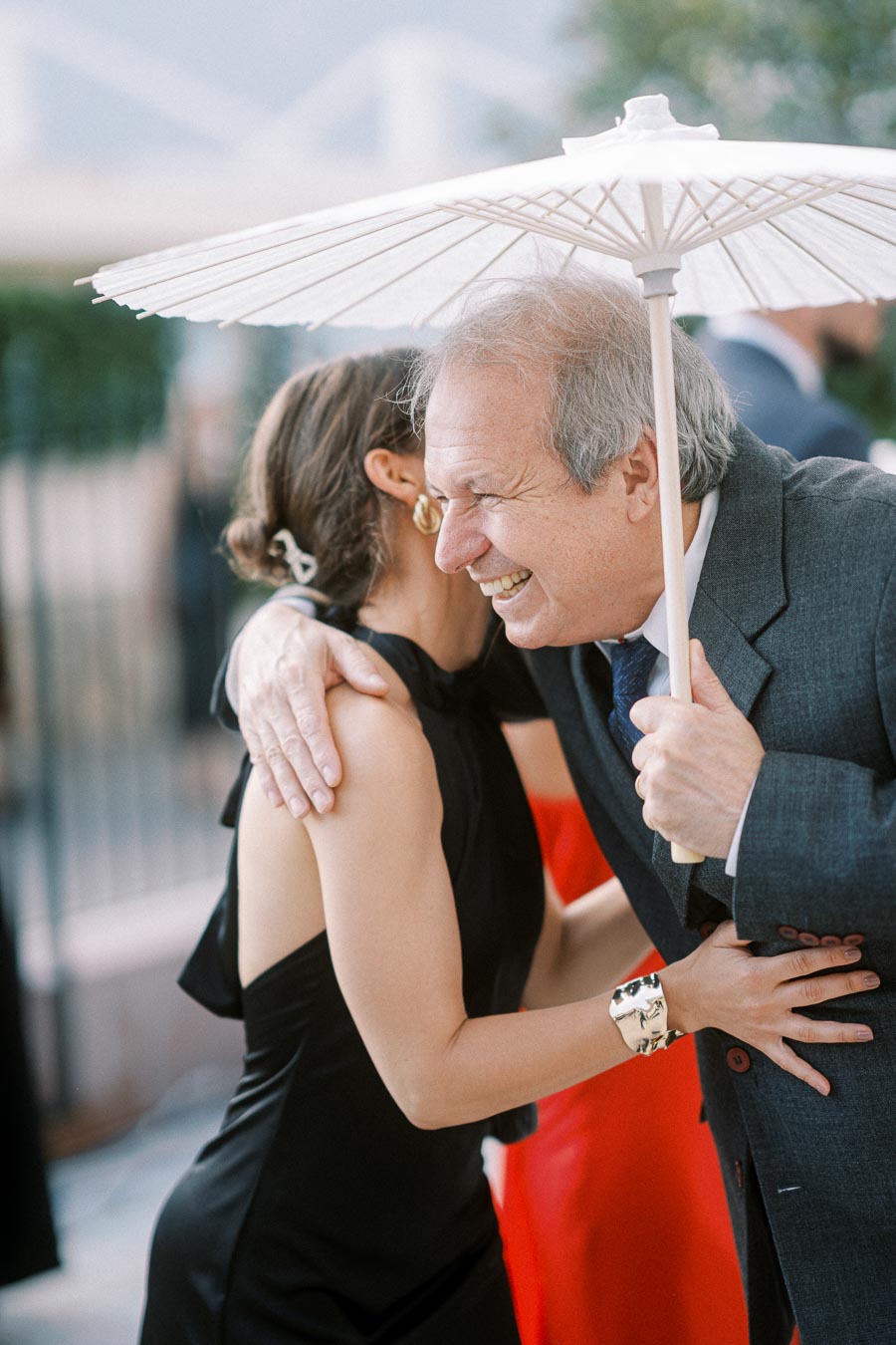 Elderly man in a suit and young woman in a black dress sharing a joyful embrace under a parasol at an outdoor event.