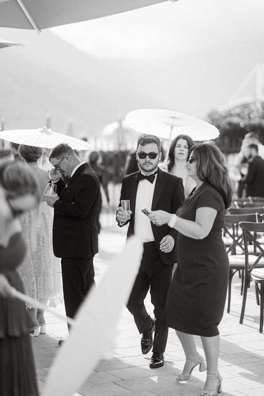 Black and white photo of elegantly dressed people at an outdoor event, featuring a man in a tuxedo and sunglasses holding a drink, with guests mingling under umbrellas in the background.