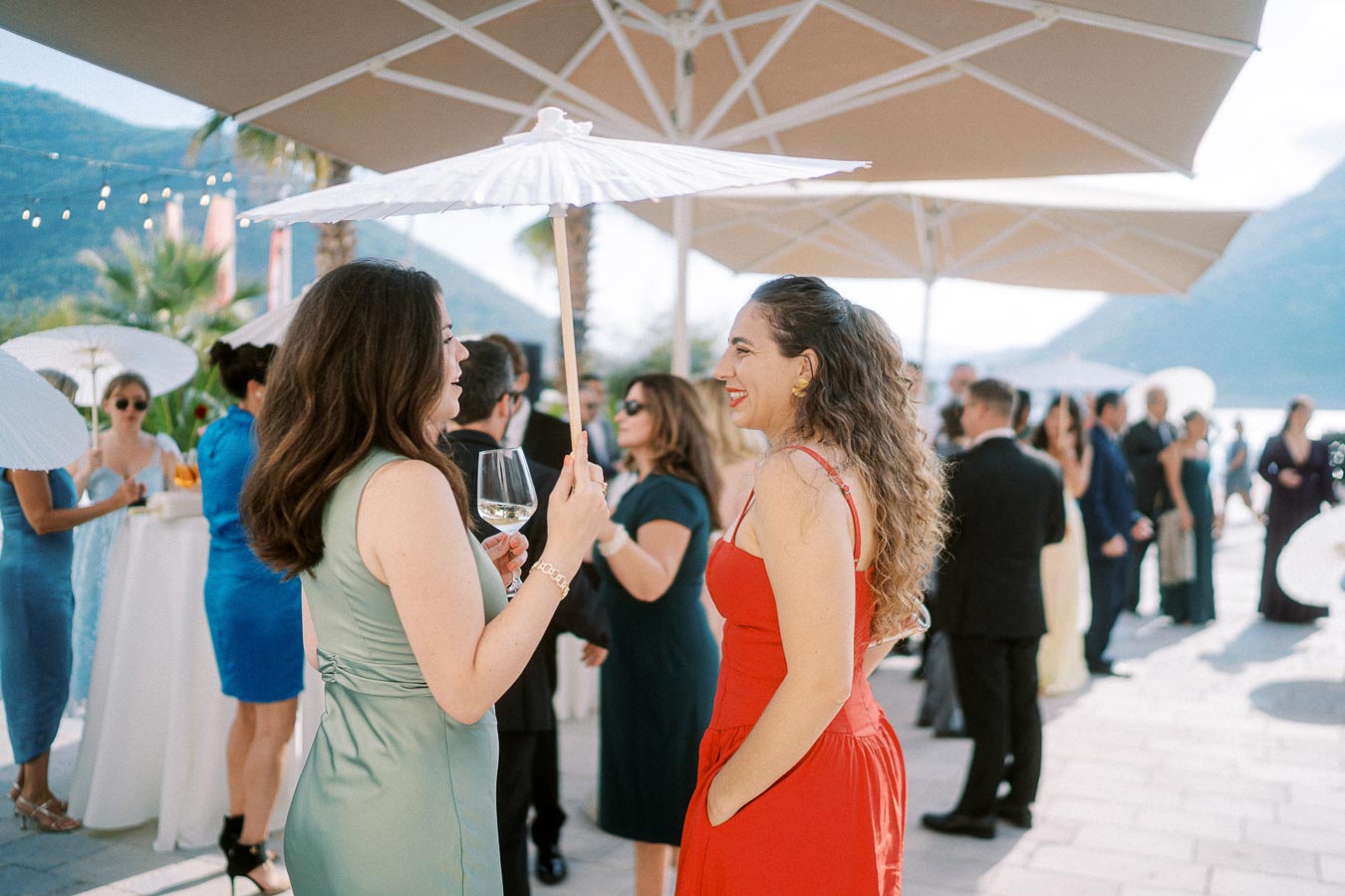 People enjoying an outdoor social event under parasols, with a scenic backdrop of mountains and water. Women in colorful dresses are chatting, one holding a wine glass.