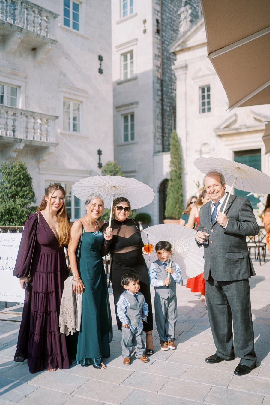 A group of well-dressed adults and children with white parasols posing on a sunny day outside a luxurious stone building, possibly at a formal event or wedding.