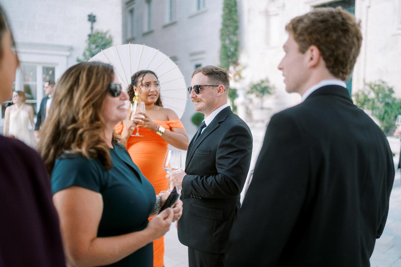 A group of elegantly dressed people socializing at an outdoor event, with a woman in an orange dress holding a parasol and a man in a black suit holding a wine glass.