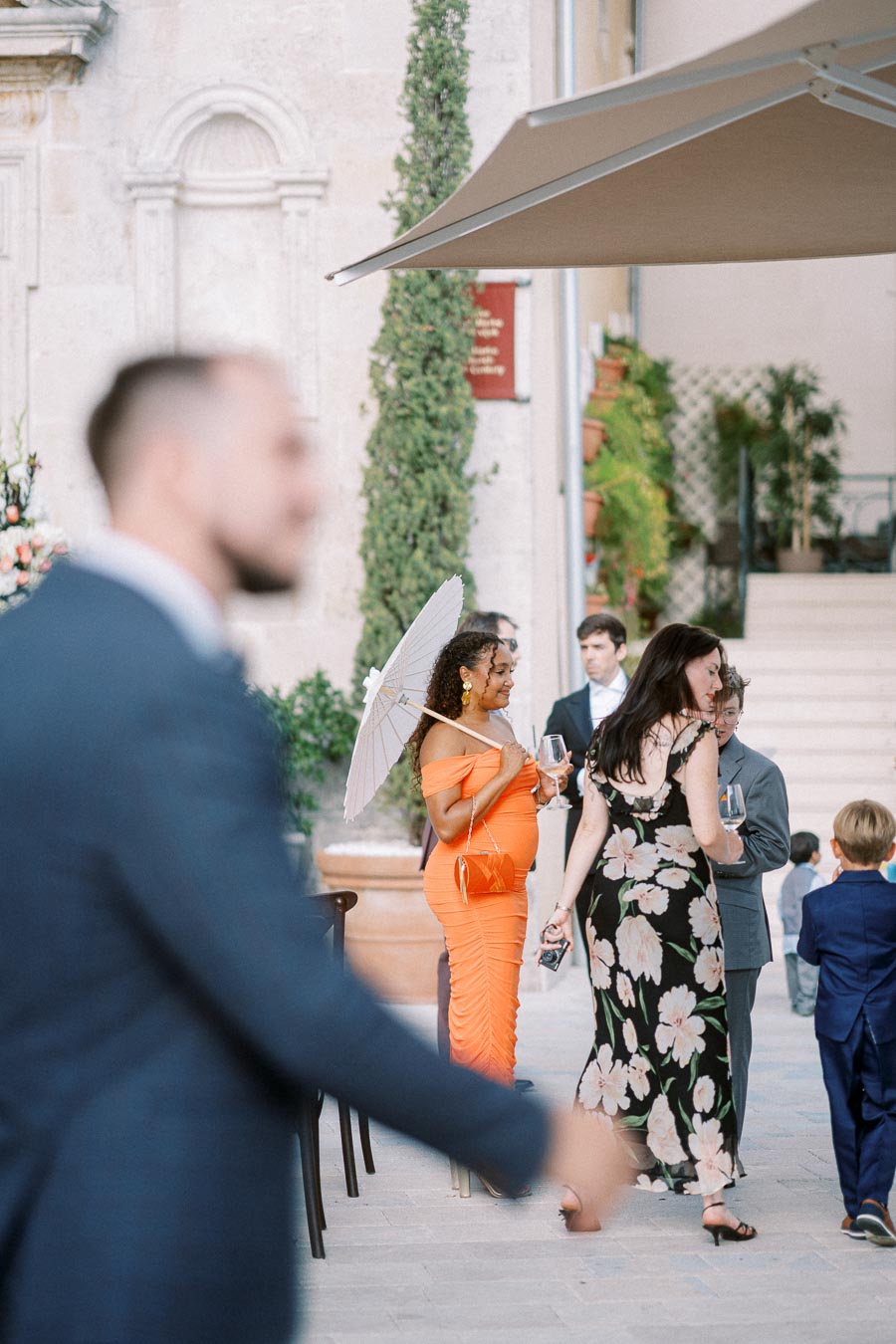 Guests mingling at an outdoor wedding reception, with a woman in an orange dress holding a parasol and a glass of wine, surrounded by elegantly dressed attendees.