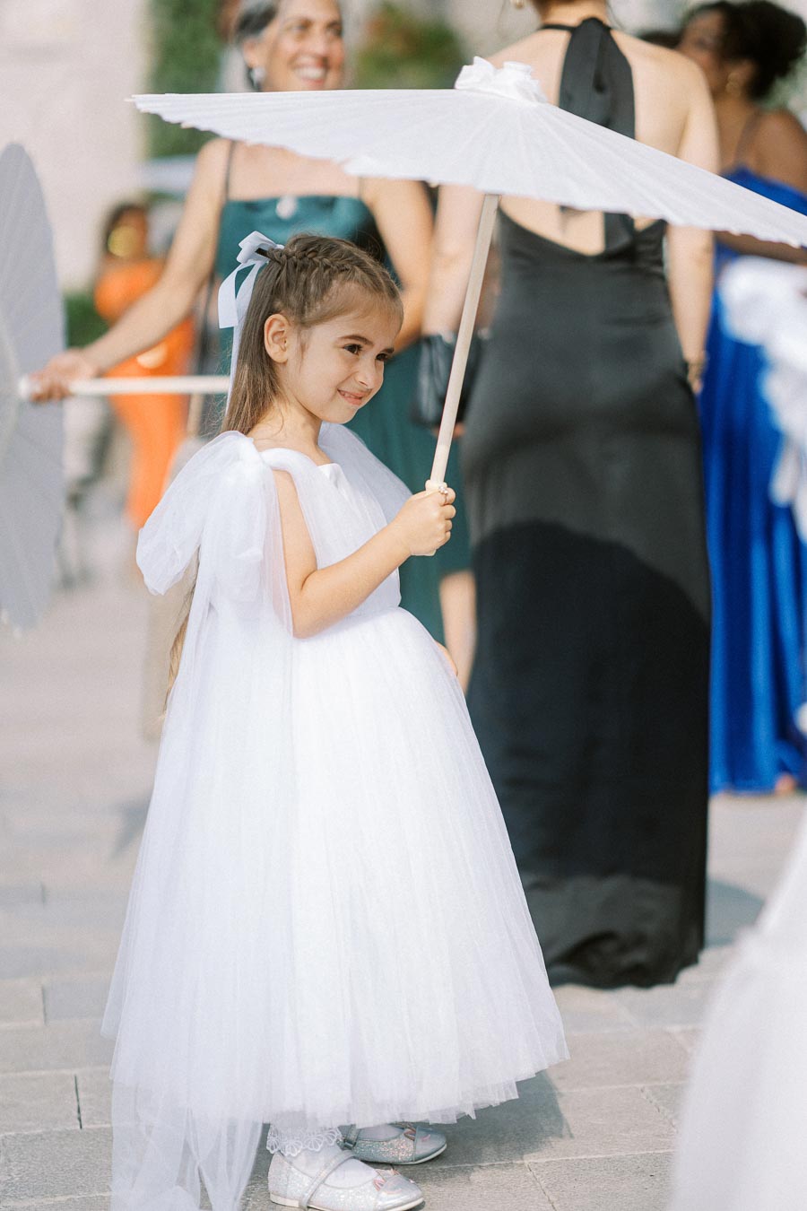 Young girl in a white dress holding a parasol at an outdoor event, surrounded by elegantly dressed people.