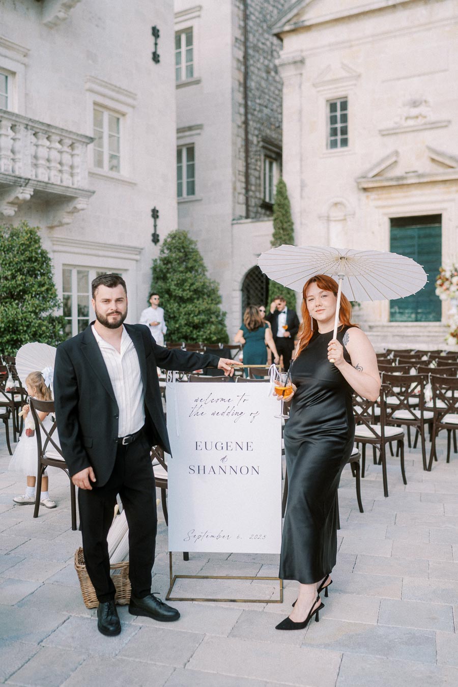 A couple poses elegantly at an outdoor wedding venue, holding a white parasol and cocktail. A welcome sign displays the names Eugene & Shannon along with the date September 6, 2023. The backdrop features a historic stone building with greenery.