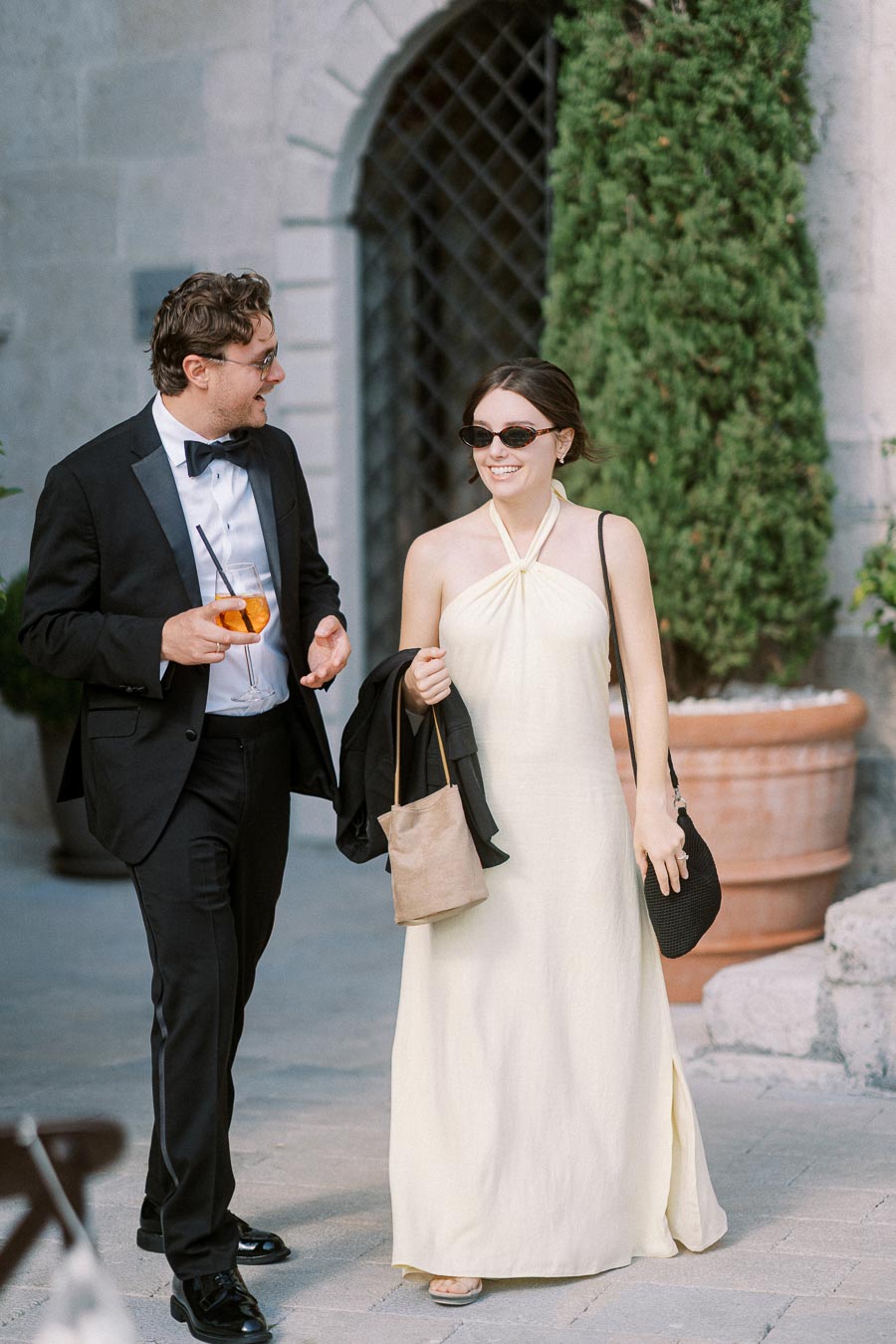 A couple dressed elegantly, walking outdoors at a formal event. The man wears a black tuxedo and holds a drink, while the woman is in a light yellow halter dress and sunglasses, carrying a jacket and a purse. They are smiling and conversing in a sophisticated garden setting.