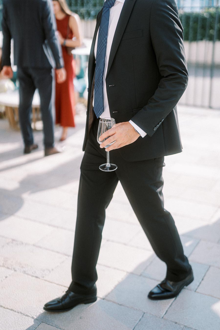A well-dressed person in a black suit holding a wine glass at an outdoor event, with other elegantly dressed attendees visible in the background.