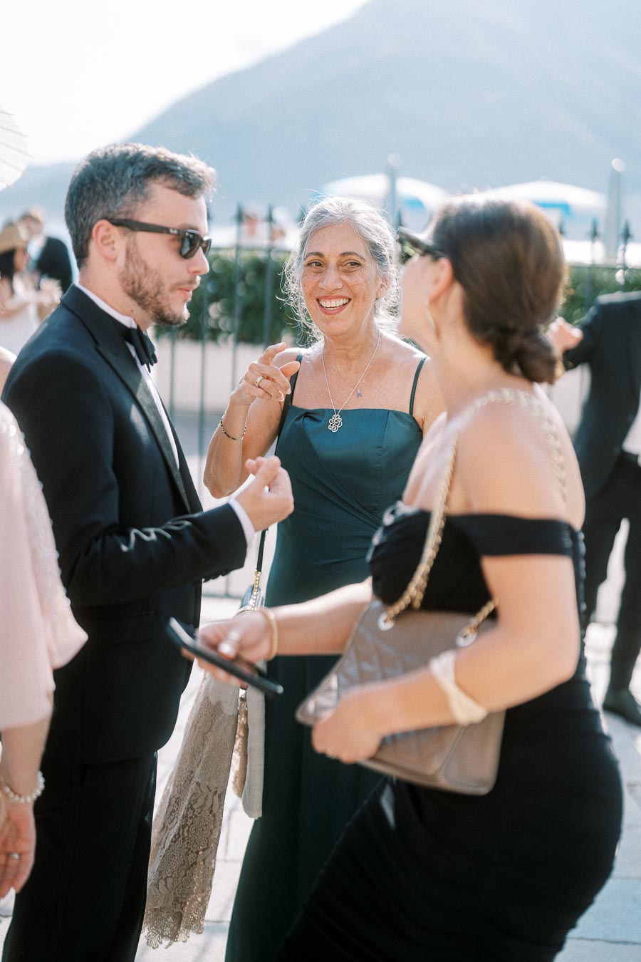 A group of elegantly dressed people socializing at an outdoor event, with a scenic mountain backdrop under a clear sky.