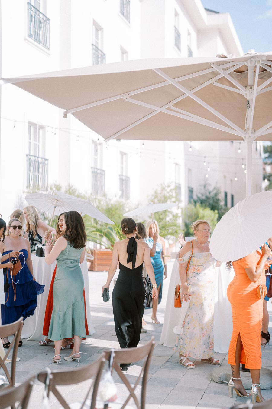 Guests at an outdoor summer event in elegant dresses, mingling under large umbrellas. Some hold white parasols, creating a festive atmosphere against a backdrop of a white building.