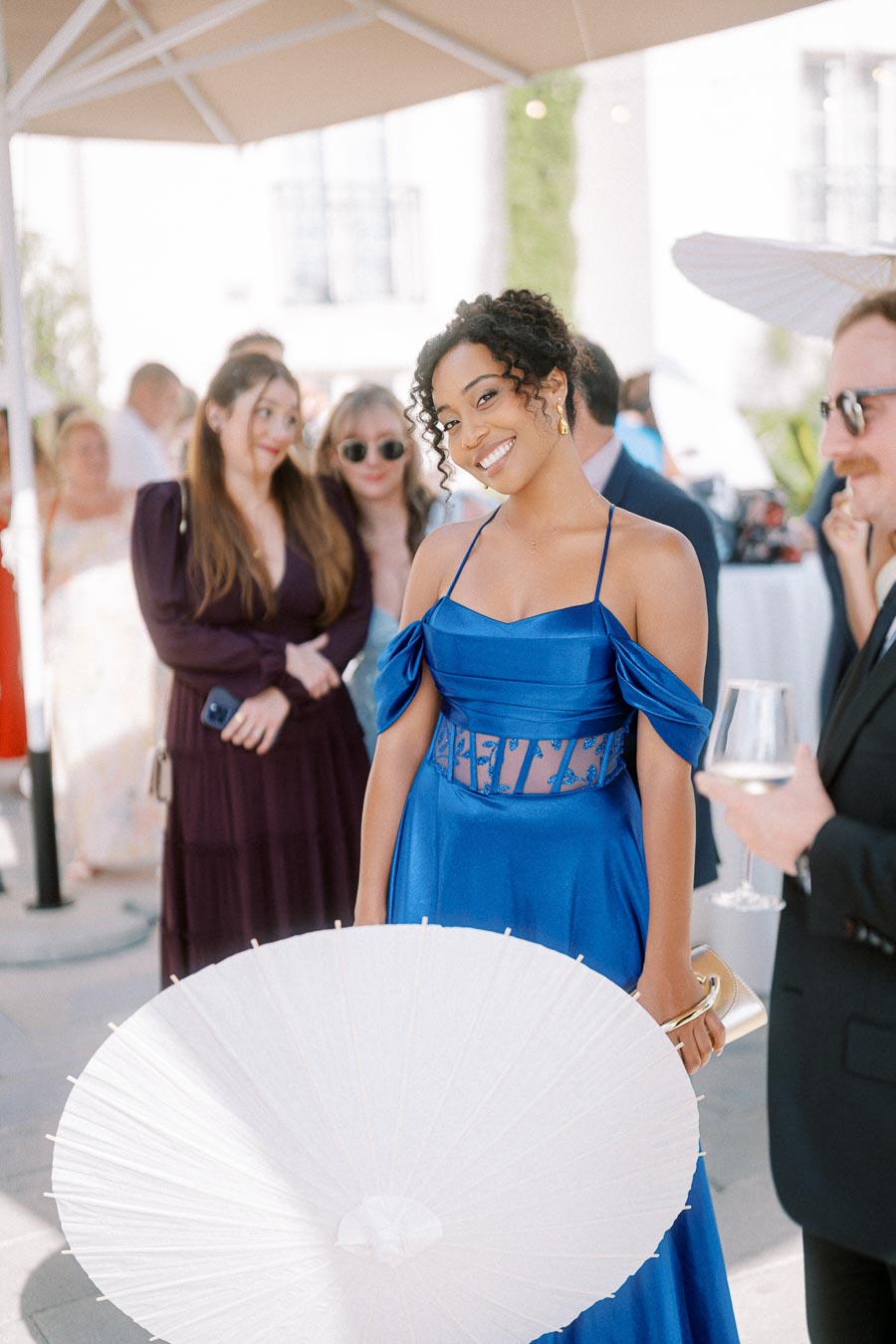 Smiling woman in a vibrant blue dress holding a white parasol at an outdoor event, surrounded by elegantly dressed guests.