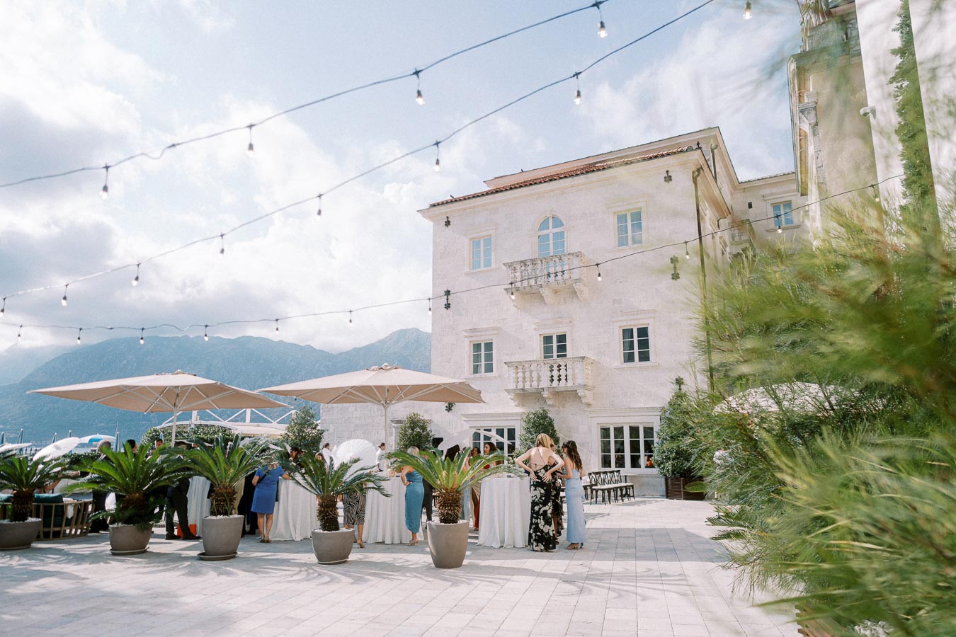 Outdoor wedding reception at a picturesque venue with string lights, white building, and mountain view in the background.