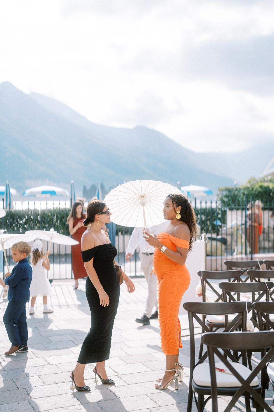 Two elegantly dressed women in vibrant dresses, one holding a white parasol, converse at an outdoor event with mountains in the background. A child in formal attire and other guests with parasols are seen in the background, creating a festive atmosphere.