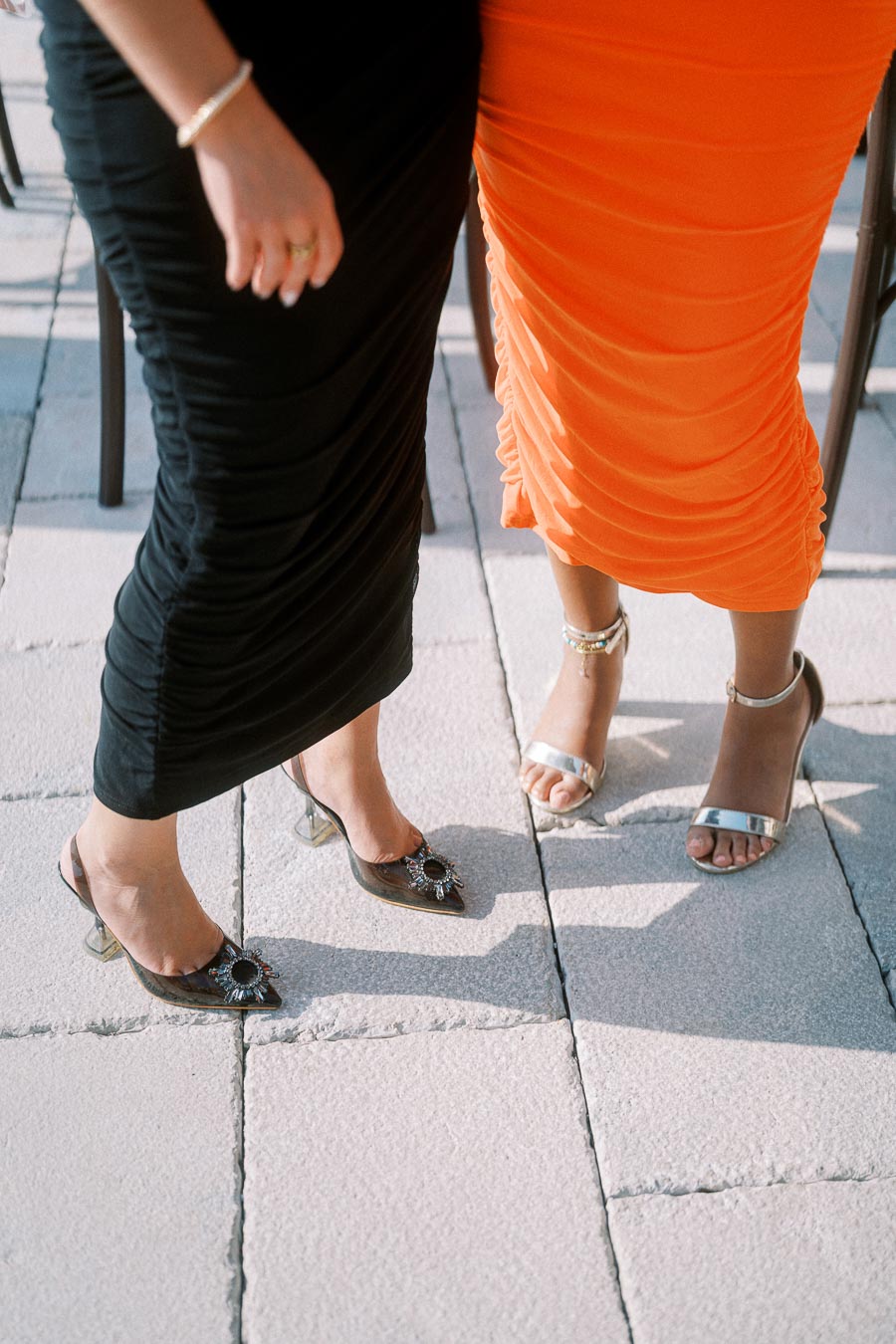 Two women in elegant evening dresses, one in black and the other in orange, showcasing fashionable high heels on a tiled surface.