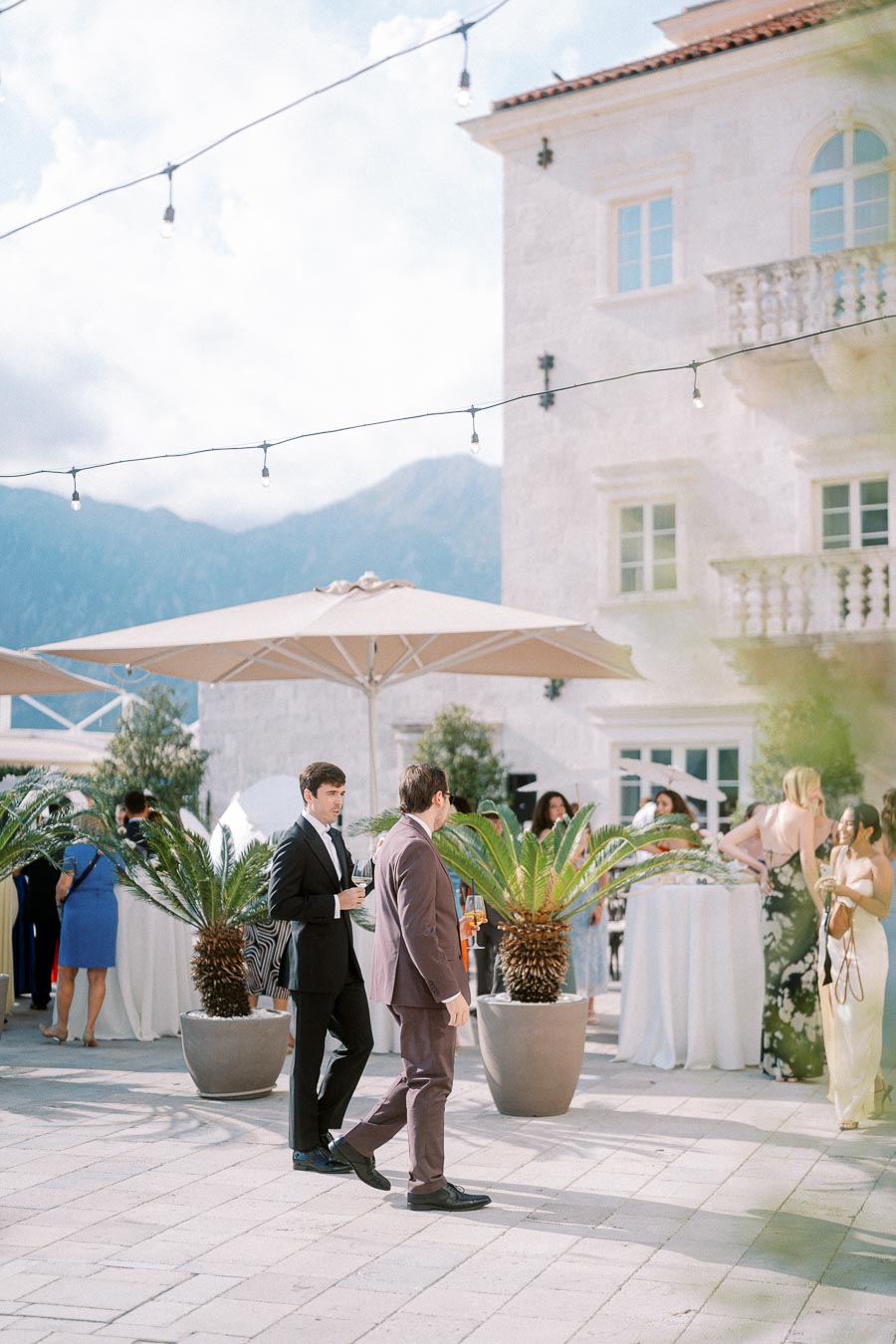 Elegant outdoor wedding reception with guests socializing under string lights, surrounded by potted palm plants and a stunning villa backdrop in the mountains.