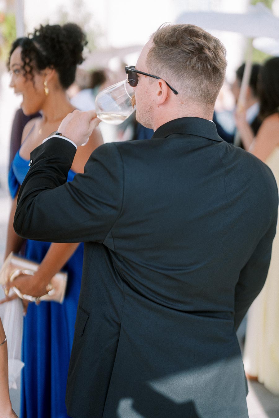 Man in a black suit and sunglasses drinking wine at an outdoor event, surrounded by elegantly dressed guests in a sunny setting.