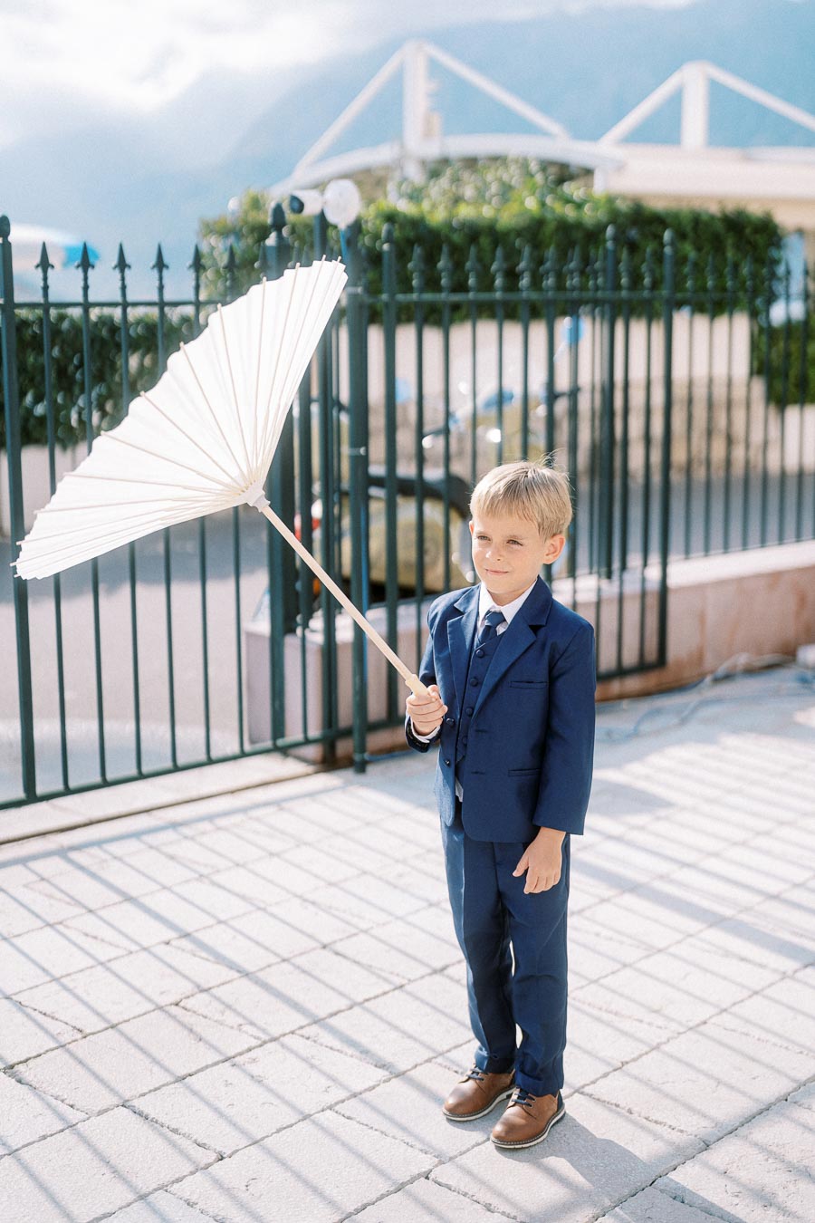 Young boy in a blue suit holding a white parasol on a sunny day, standing in front of a wrought iron fence.