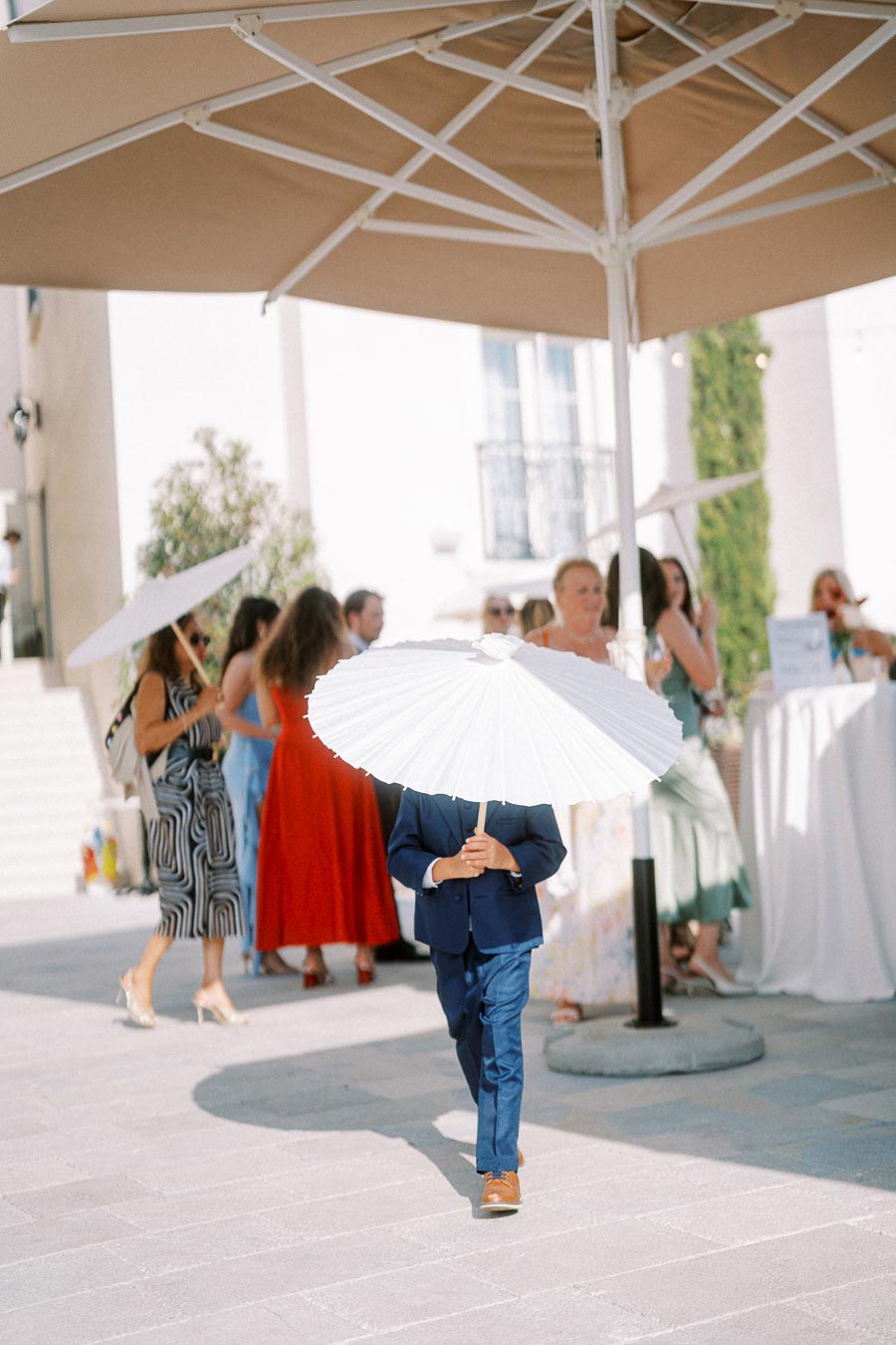 Child in a suit holding a white parasol at an outdoor event, surrounded by elegantly dressed people and sunshades, capturing a festive and stylish atmosphere.