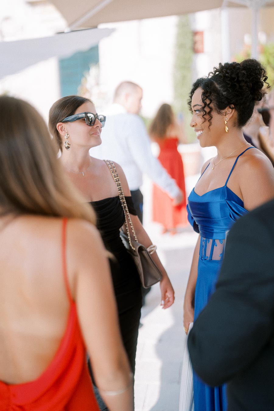 A group of elegantly dressed people engaged in conversation at a social gathering, with women wearing stylish dresses and accessories, creating a lively and fashionable atmosphere.
