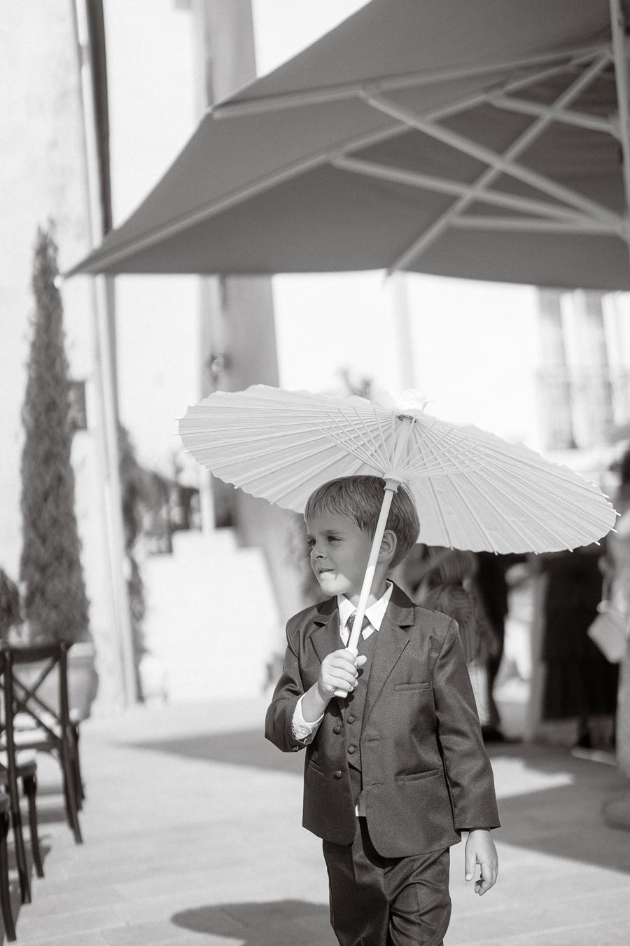 Black and white photo of a young boy in a suit holding a paper umbrella, walking outdoors on a sunny day.
