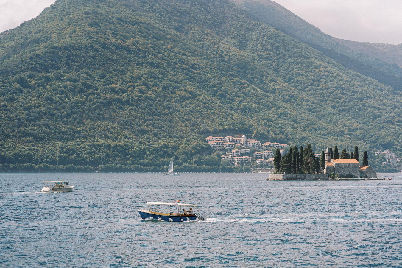 A scenic view of a calm bay with boats gliding on the blue water, an island with a historic building surrounded by cypress trees, and lush green mountains in the background under a partially cloudy sky.