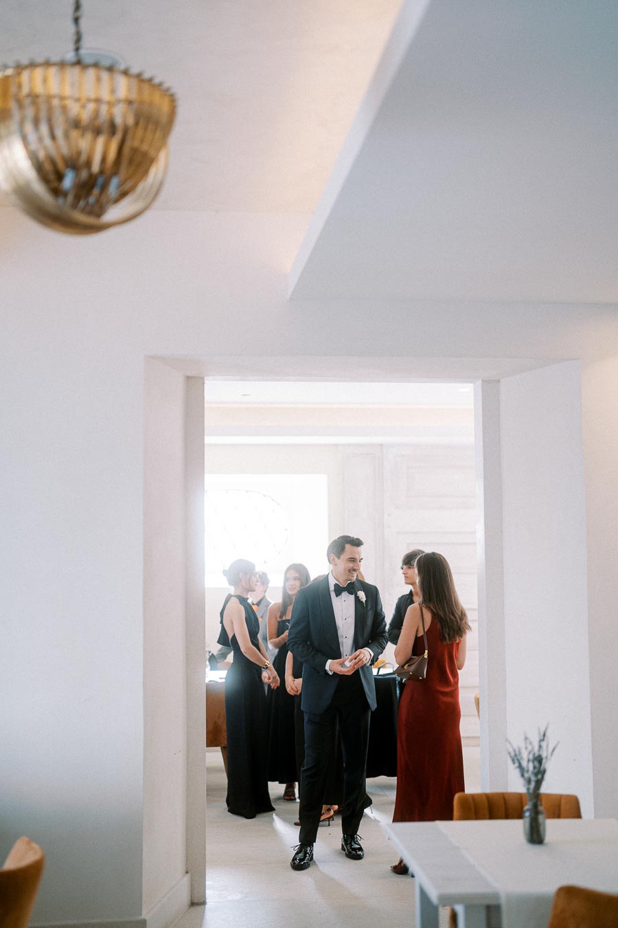 Elegant gathering at a formal event with people in stylish attire, including a man in a tuxedo and women in evening dresses, engaging in conversation inside a modern, minimalist room with a decorative chandelier.