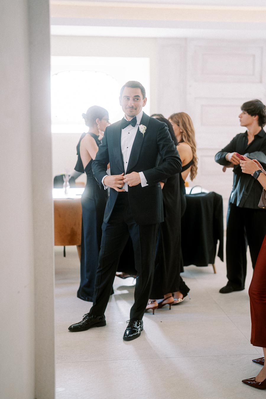A man in a black tuxedo standing confidently in an elegant indoor setting, surrounded by people in semi-formal attire, preparing for a formal event or celebration.