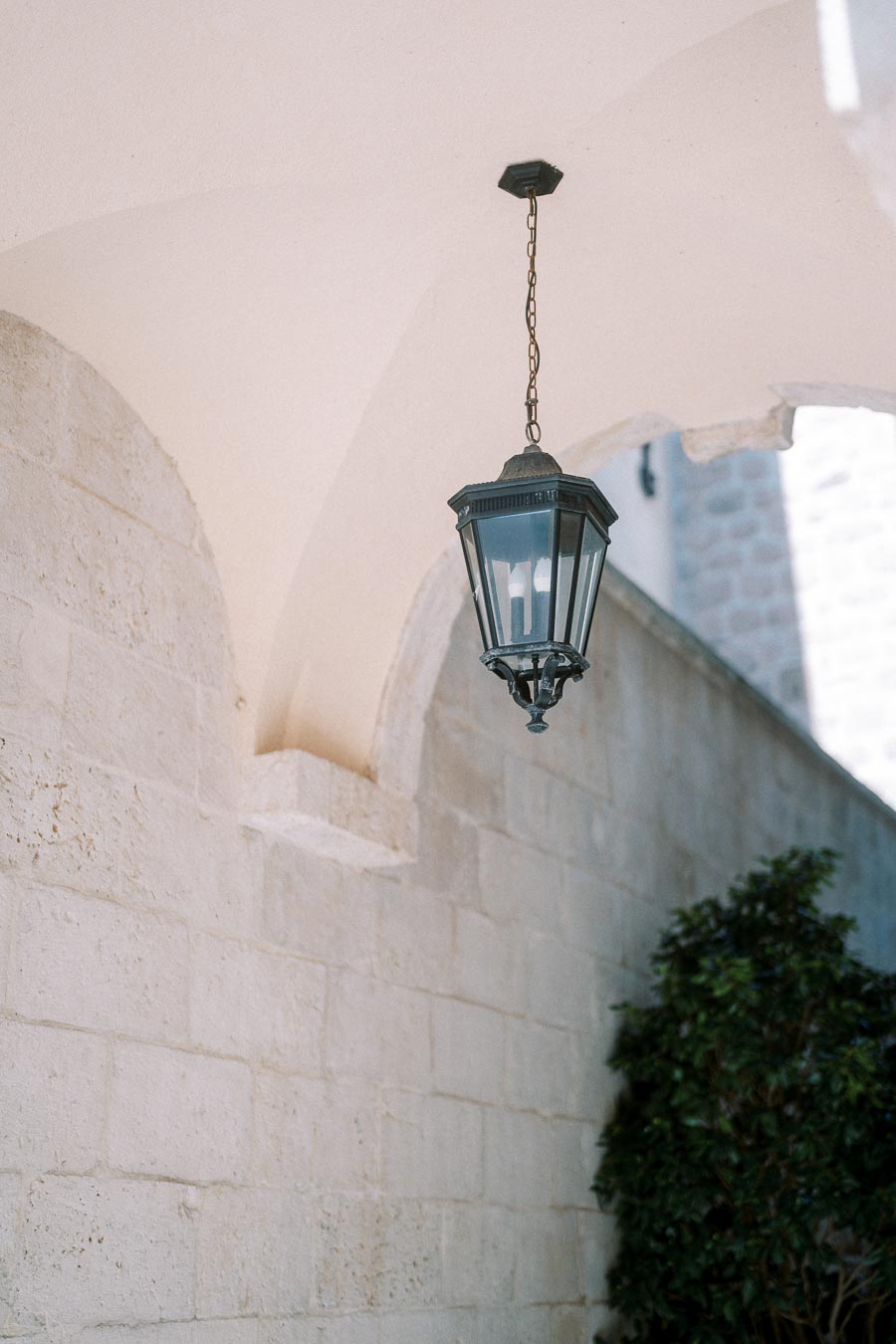 Elegant black lantern hanging from an arched outdoor ceiling, casting soft light on a stone wall with green foliage nearby.
