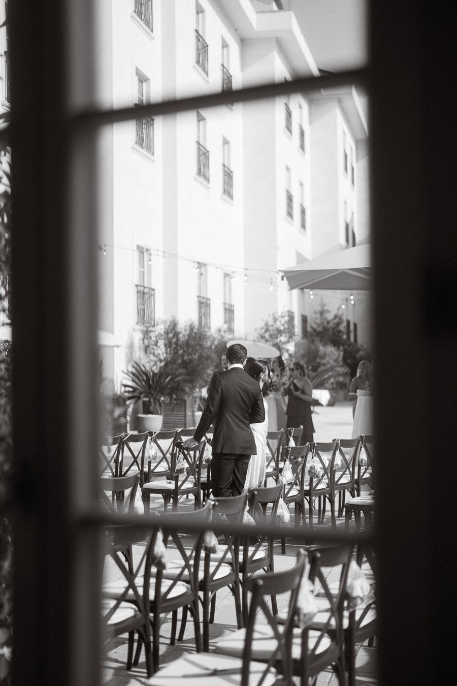 Black and white photo of an outdoor wedding ceremony, featuring a couple standing among empty chairs, framed through a window with a view of a historic building in the background.