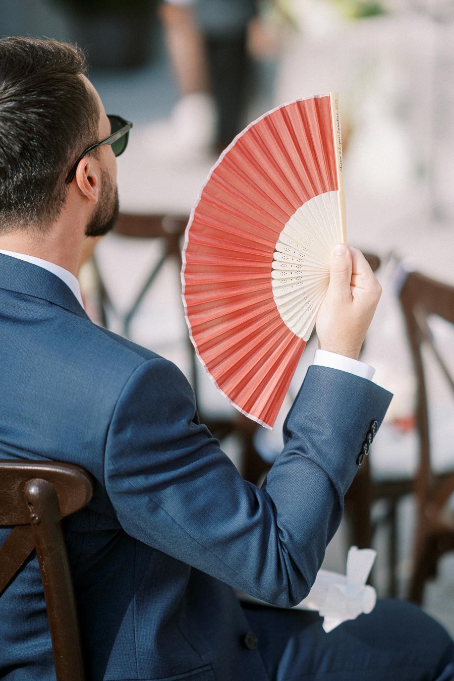 Man in a blue suit sitting on a chair, holding a red and white paper fan, possibly at an outdoor event.
