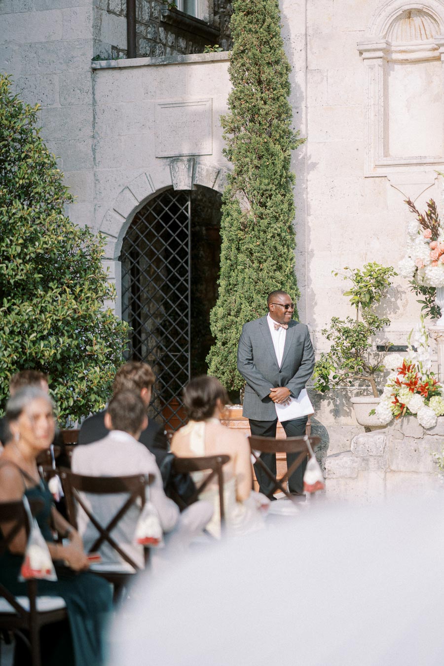 Outdoor wedding ceremony with a smiling officiant in a suit standing in front of an elegant stone wall and greenery, surrounded by seated guests.