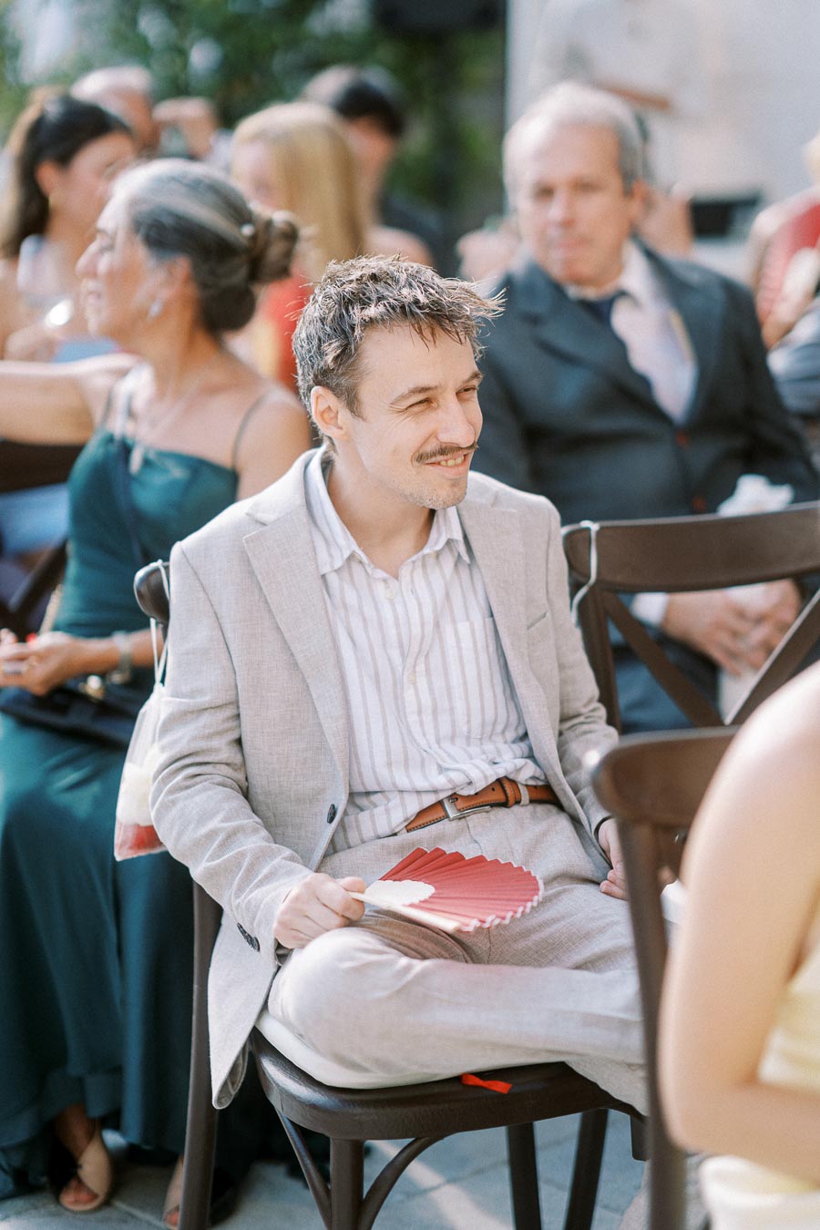 A man in a light gray suit sitting on a chair outdoors at an event, smiling and holding a red fan, with other attendees in the background.