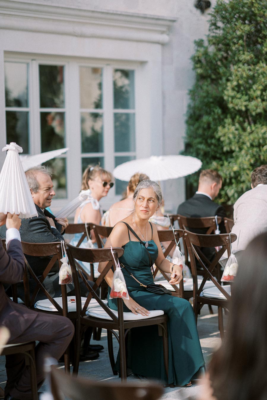 Elegant outdoor wedding guest seated on a wooden chair, wearing a teal dress and surrounded by others holding white parasols, with a backdrop of lush greenery and a classic building facade.