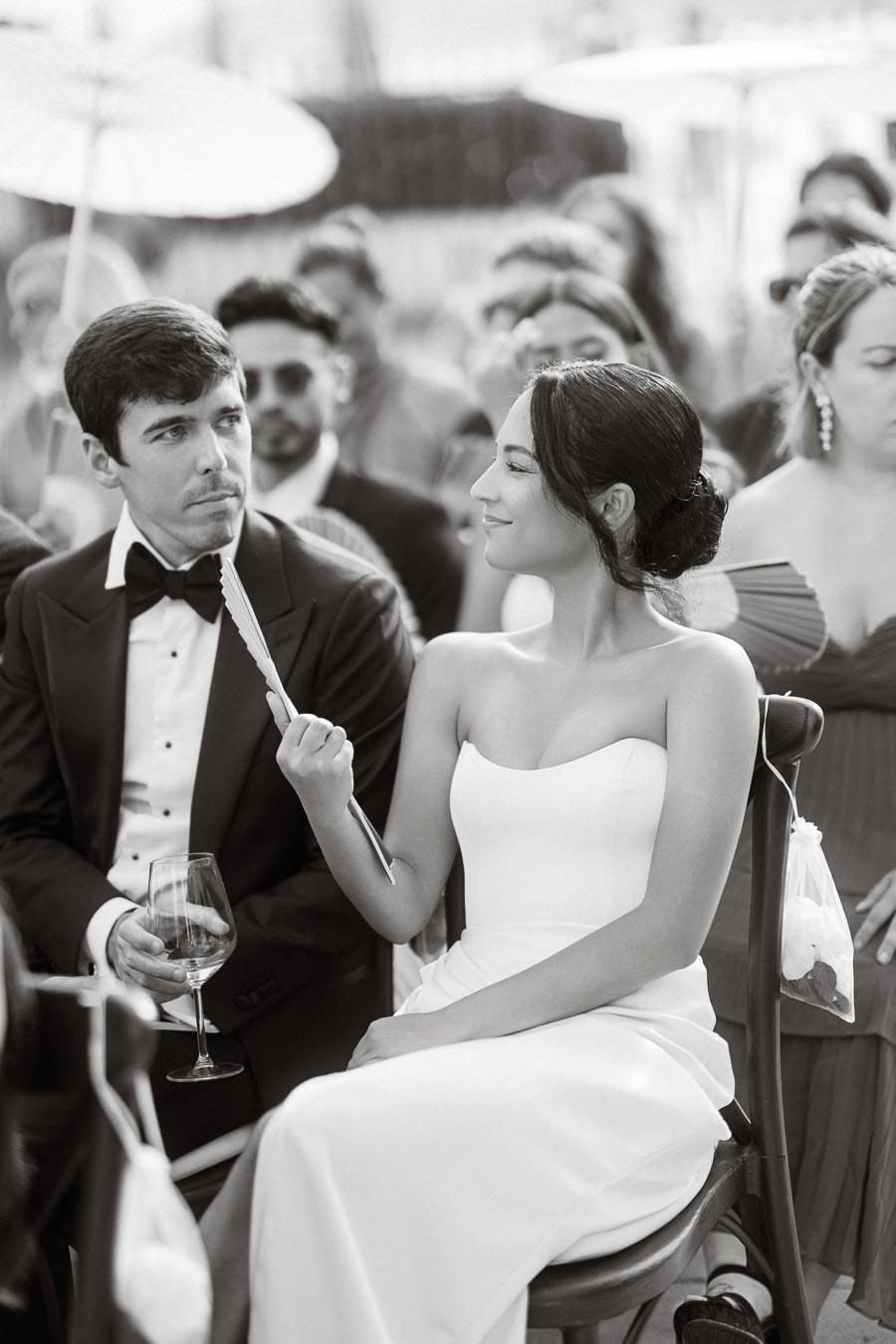 Black and white image of a bride and groom sitting together, with the bride playfully holding a fan and smiling. She is wearing an elegant strapless wedding gown, while the groom is dressed in a classic black tuxedo, holding a wine glass, surrounded by wedding guests.